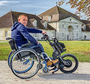 Homme en fauteuil roulant avec une roue électrique BATEC devant une maison.